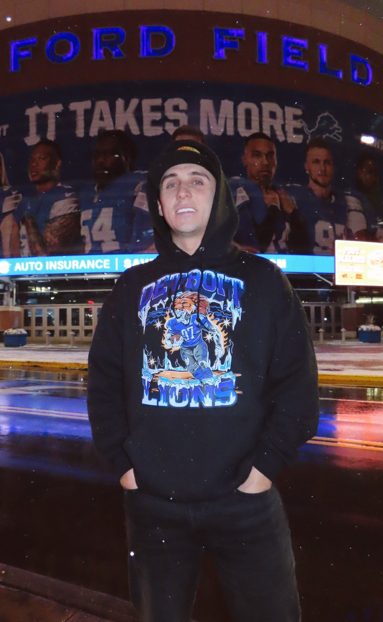 Person wearing a Detroit Lions hoodie in front of a large screen displaying football players and 'Ford Field' text.